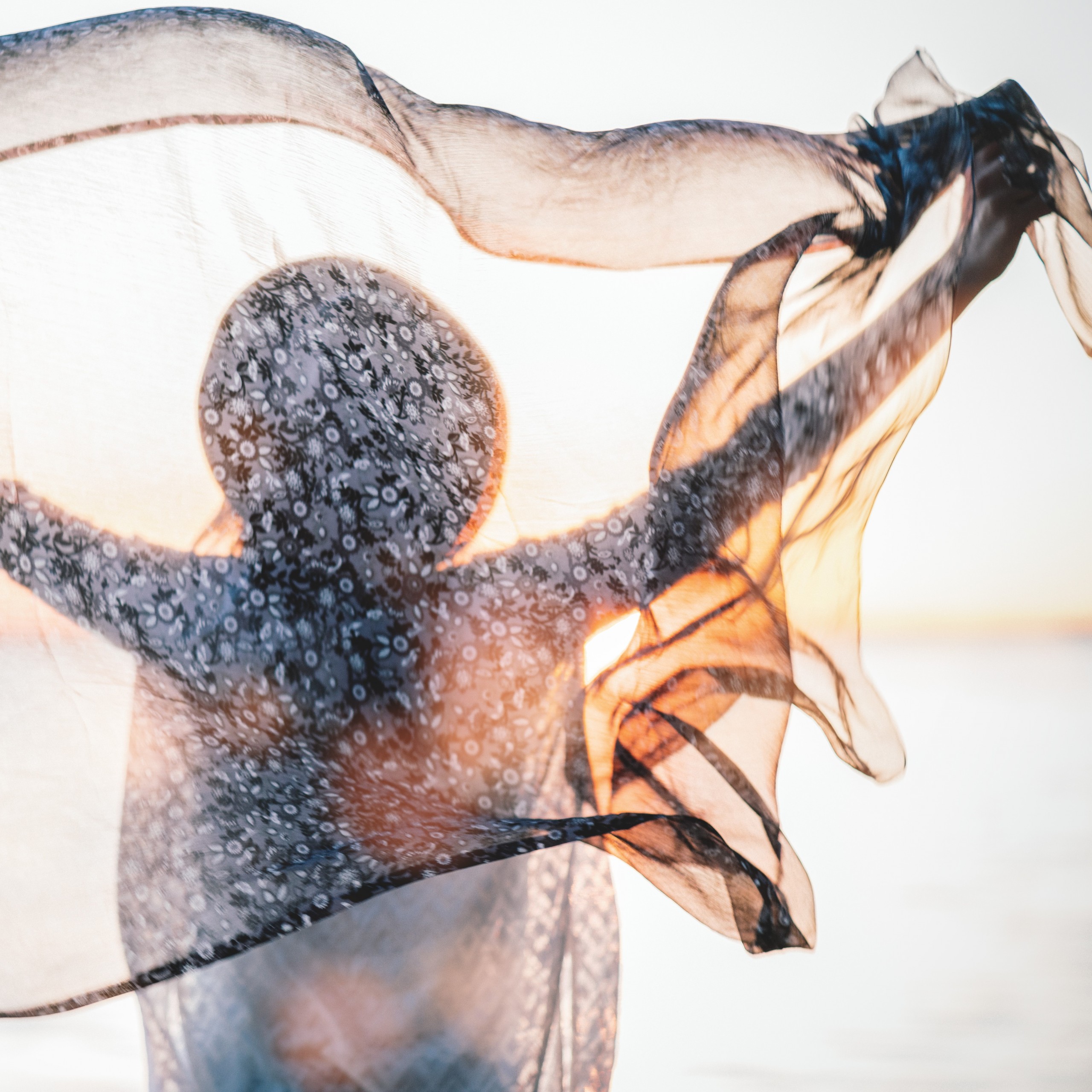 Beautiful boho girl with her hands raised up holds a silk scarf on breeze. Handsome woman look on evening sun on horizon
